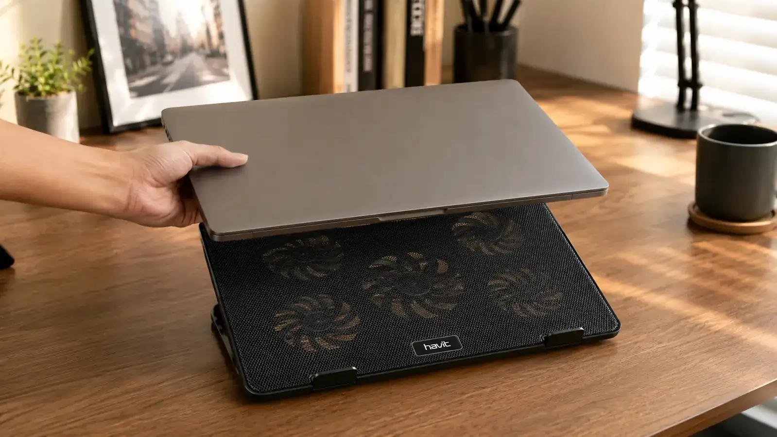 Hand sliding a laptop onto a slim mesh cooling pad on a wooden desk