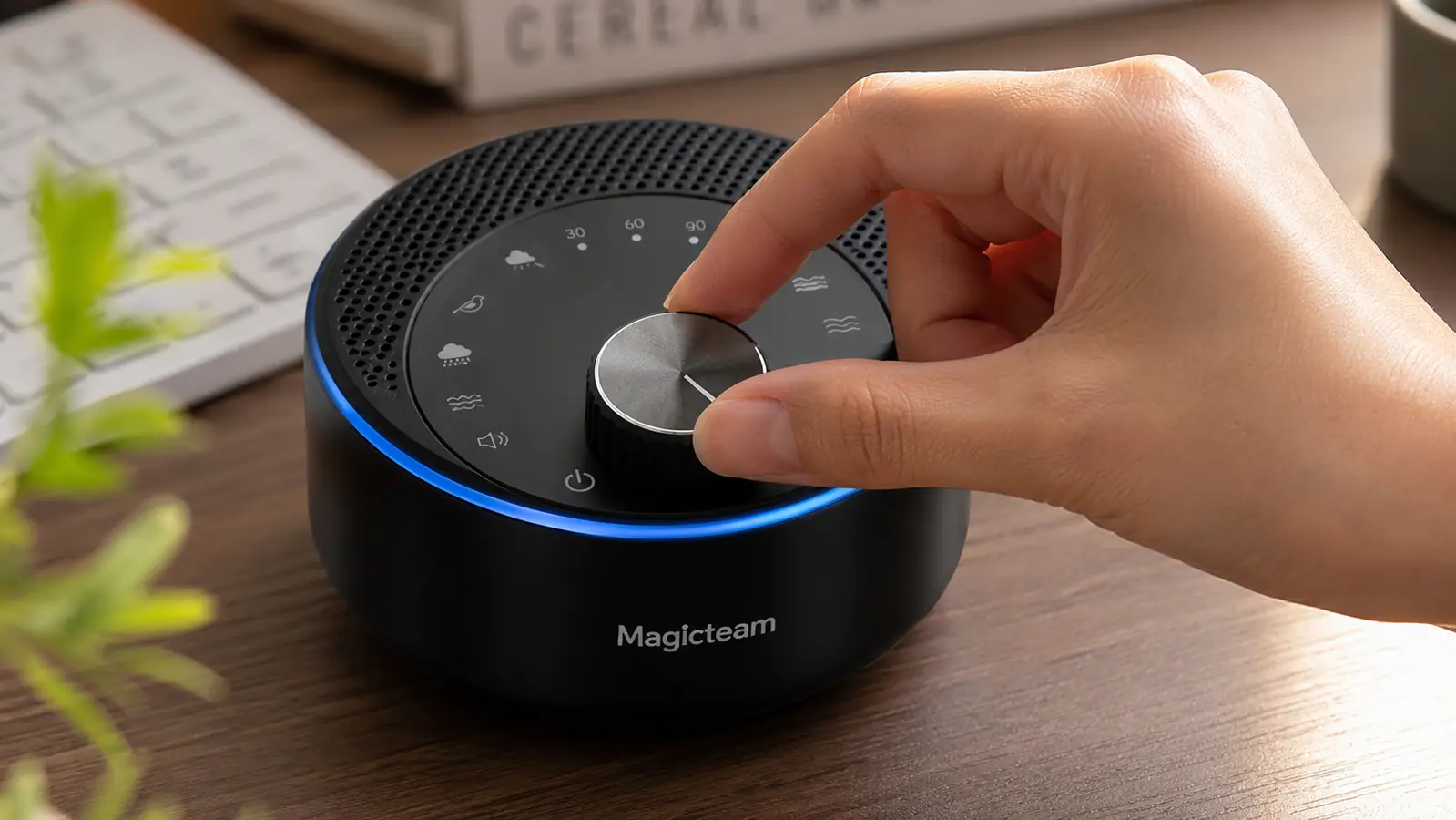 Hand adjusting the volume dial on a white noise machine positioned on a desk next to a keyboard