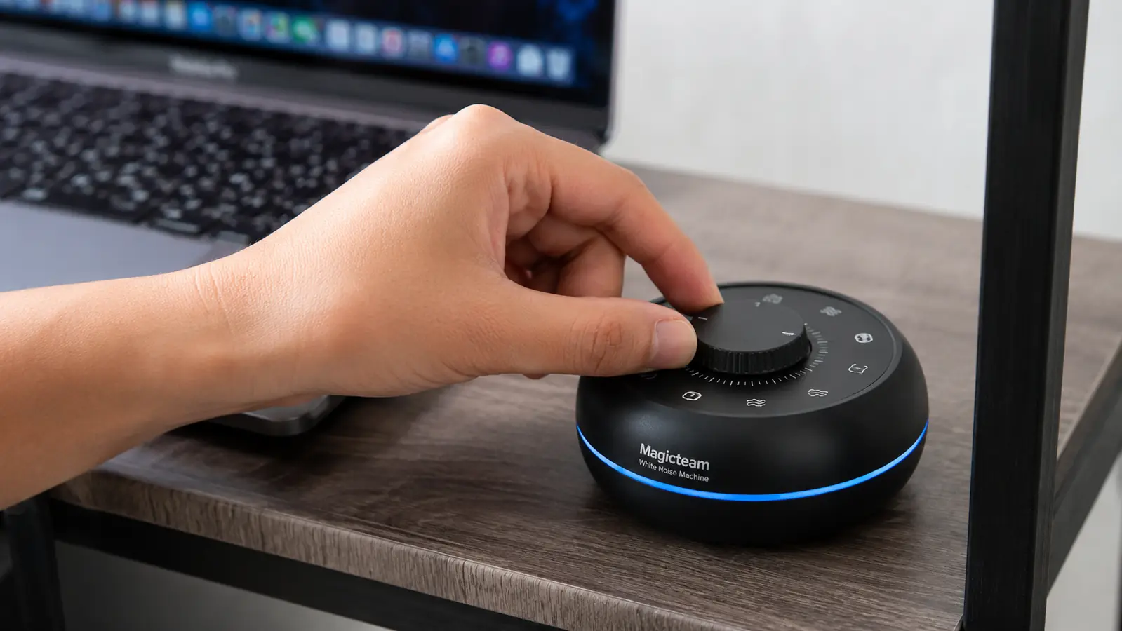 Hand adjusting the volume dial on a Magicteam white noise machine sitting on a desk shelf